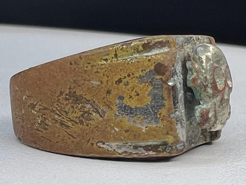 Puede incluir: Un anillo desgastado de color bronce con un dise&ntilde;o detallado, posiblemente antiguo, en la cara. El anillo muestra signos de edad y desgaste, con una apariencia deslustrada y textura visible. El dise&ntilde;o parece ser un relieve.