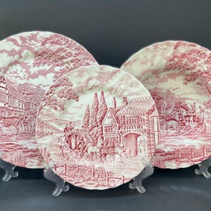 May include: Three pink and white ceramic plates with a cottage and horse-drawn carriage design. The plates are on clear stands.