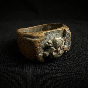 Puede incluir: Un anillo de bronce envejecido con una banda rectangular y un dise&ntilde;o esculpido detallado en la parte delantera. El dise&ntilde;o presenta un motivo de calavera y huesos cruzados. El anillo tiene una apariencia desgastada y antigua.