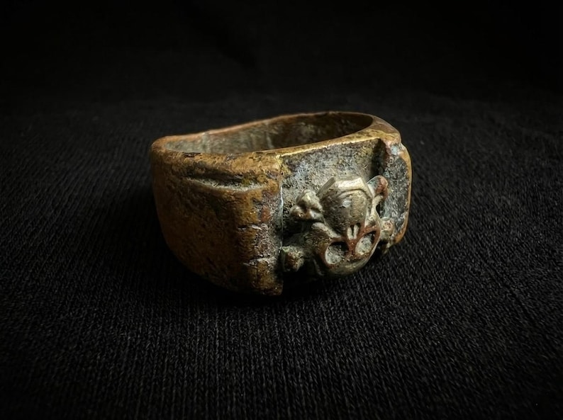 Puede incluir: Un anillo de bronce envejecido con una banda rectangular y un dise&ntilde;o esculpido detallado en la parte delantera. El dise&ntilde;o presenta un motivo de calavera y huesos cruzados. El anillo tiene una apariencia desgastada y antigua.