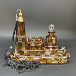 May include: An amber-colored vanity set with a perfume bottle, jars, and a tray. The perfume bottle has a gold-colored atomizer and a black tassel. The set is made of cut glass with a geometric pattern.