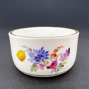 Pode incluir: Uma pequena xícara de porcelana branca com uma borda dourada e um desenho floral. As flores são pintadas em tons de amarelo, laranja, rosa, roxo e azul.