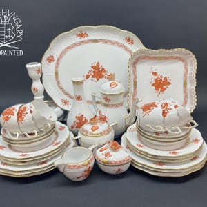 May include: A Herend Hungary handpainted porcelain dinnerware set featuring white pieces with orange floral patterns and gold trim. The collection includes plates, cups, a teapot, a sugar bowl, a creamer, a vase, and a candlestick with scalloped edges.
