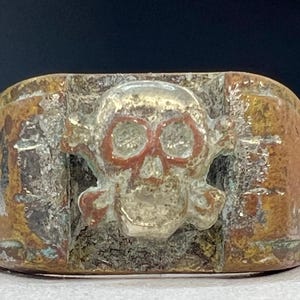 Puede incluir: Anillo de metal envejecido con un dise&ntilde;o de calavera y huesos cruzados. Los cuencas de los ojos de la calavera est&aacute;n resaltadas en rojo. El anillo tiene un aspecto desgastado con tonos marrones, plateados y cobrizos.