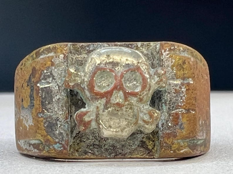 Puede incluir: Anillo de metal envejecido con un dise&ntilde;o de calavera y huesos cruzados. Los cuencas de los ojos de la calavera est&aacute;n resaltadas en rojo. El anillo tiene un aspecto desgastado con tonos marrones, plateados y cobrizos.