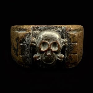 Puede incluir: Anillo de bronce con un dise&ntilde;o de calavera y huesos cruzados. La calavera tiene cuencas de ojos y mand&iacute;bula detalladas, enmarcada en un marco rectangular. El anillo tiene una apariencia desgastada, posiblemente antigua.