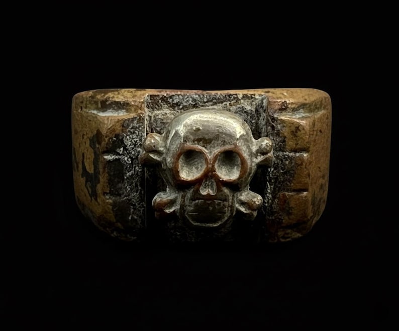 Puede incluir: Anillo de bronce con un dise&ntilde;o de calavera y huesos cruzados. La calavera tiene cuencas de ojos y mand&iacute;bula detalladas, enmarcada en un marco rectangular. El anillo tiene una apariencia desgastada, posiblemente antigua.