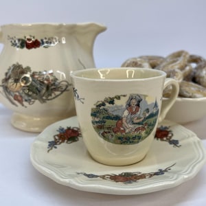Puede incluir: Un juego de taza y platillo de porcelana blanca con un diseño floral y una escena de una mujer con una cesta de flores. El juego está al lado de una jarra de crema de porcelana blanca con un diseño floral.