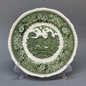 Może przedstawiać: Biało-zielony talerz dekoracyjny z falistym brzegiem. Talerz przedstawia zieloną scenę krajobrazową z zamkiem, drzewami i końmi. Obramowanie zdobią zielone wzory kwiatowe. Talerz jest prezentowany na przezroczystym stojaku.