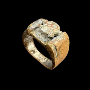 Puede incluir: Un anillo desgastado, de tono bronce, con un dise&ntilde;o de tortuga. El anillo presenta una cara rectangular con una figura de tortuga en relieve y una banda con un aspecto desgastado y texturizado. El anillo est&aacute; sobre un fondo negro.