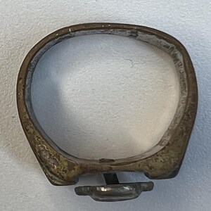 Puede incluir: Un anillo antiguo con una banda de color bronce deslustrado. El anillo presenta un peque&ntilde;o engaste rectangular con una piedra oscura, posiblemente negra. La banda tiene una forma ligeramente irregular, lo que sugiere que puede ser hecho a mano o vintage.