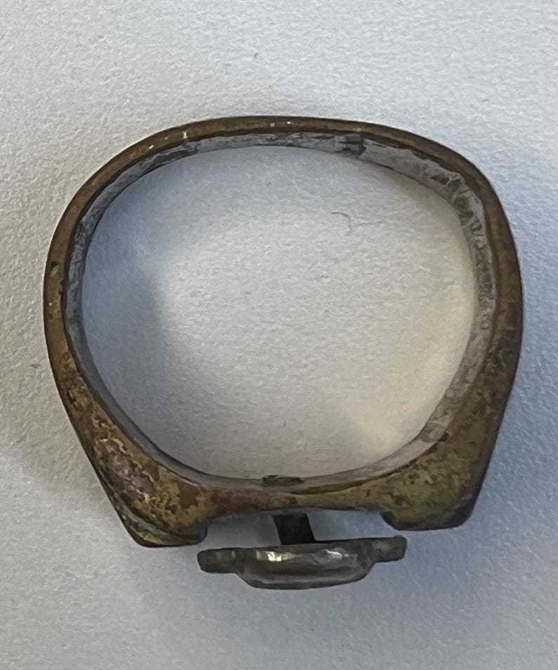 Puede incluir: Un anillo antiguo con una banda de color bronce deslustrado. El anillo presenta un peque&ntilde;o engaste rectangular con una piedra oscura, posiblemente negra. La banda tiene una forma ligeramente irregular, lo que sugiere que puede ser hecho a mano o vintage.