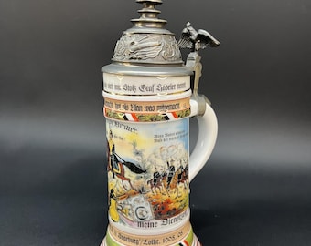 May include: A decorative ceramic beer stein with a detailed historical scene and text. The lid features a mounted rider figurine. The base has red, white, and green bands. The stein is approximately 25 cm tall.