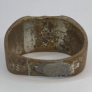 Puede incluir: Un anillo desgastado, de color bronce, con una cara rectangular. El anillo muestra signos de desgaste y edad, con &aacute;reas de decoloraci&oacute;n y una textura ligeramente &aacute;spera. El interior del anillo es visible, revelando su construcci&oacute;n hueca.