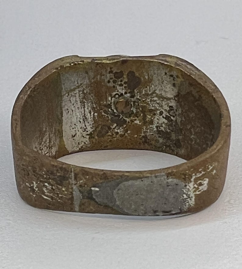 Puede incluir: Un anillo desgastado, de color bronce, con una cara rectangular. El anillo muestra signos de desgaste y edad, con &aacute;reas de decoloraci&oacute;n y una textura ligeramente &aacute;spera. El interior del anillo es visible, revelando su construcci&oacute;n hueca.