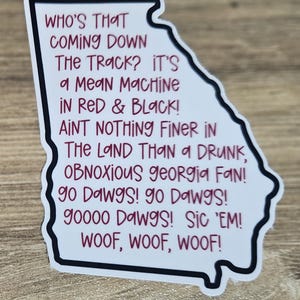 May include: A white sticker in the shape of the state of Georgia with a black border. The sticker has text in red and black, including phrases like "Mean Machine" and "Go Dawgs!" The sticker is a fan item.