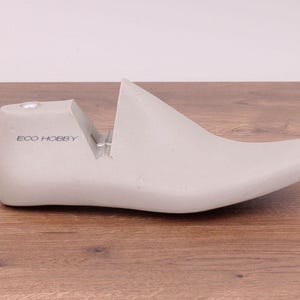 May include: A light gray shoe last with the text "ECO HOBBY" printed on the heel. The shoe form is made of a solid material and is designed to shape shoes. The last is placed on a wooden surface.