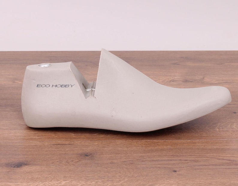 May include: A light gray shoe last with the text "ECO HOBBY" printed on the heel. The shoe form is made of a solid material and is designed to shape shoes. The last is placed on a wooden surface.