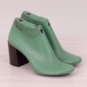 May include: Two green shoe lasts with dark brown block heels. The shoe forms are made of a solid material and have a smooth, curved design. The heels are rectangular and provide a stable base for the shoe form.