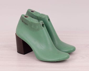 May include: Pair of green shoe lasts with dark brown block heels. The shoe forms are made of a solid material and have a smooth, curved design. The heels are rectangular and provide a stable base for the shoe form.
