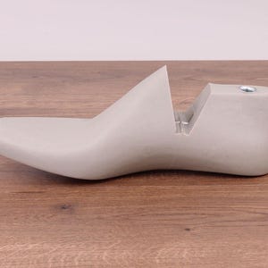 May include: A light gray shoe last, a form used for shaping shoes. The last is made of a solid material and has a smooth, curved design. It sits on a wooden surface.