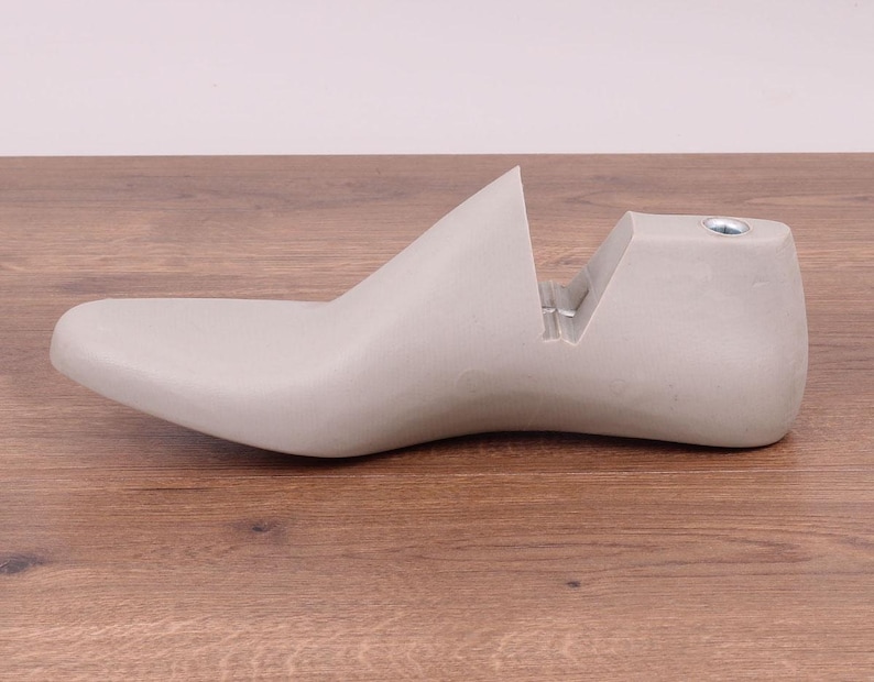 May include: A light gray shoe last, a form used for shaping shoes. The last is made of a solid material and has a smooth, curved design. It sits on a wooden surface.