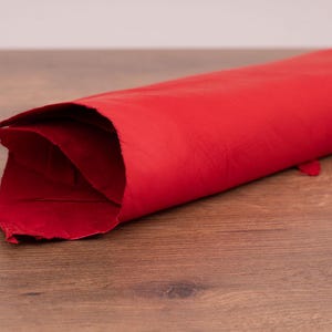 Pode incluir: Uma peça de couro vermelho vibrante enrolada com um acabamento liso e mate. O material está dobrado na borda, revelando a textura e a espessura da pele. A superfície mostra variações naturais e uma cor vermelha ousada e consistente.