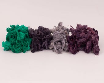 May include: Four distinct piles of loose, textured fiber arranged in a horizontal row. The colors from left to right are a vibrant emerald green, a deep charcoal black, a soft heather gray, and a rich, dark plum purple, all with a fuzzy appearance.