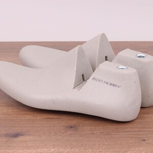 May include: Two light gray shoe lasts, one with the text "ECO HOBBY" printed on the side, are displayed on a wooden surface. The lasts are designed to maintain the shape of shoes during storage or repair.