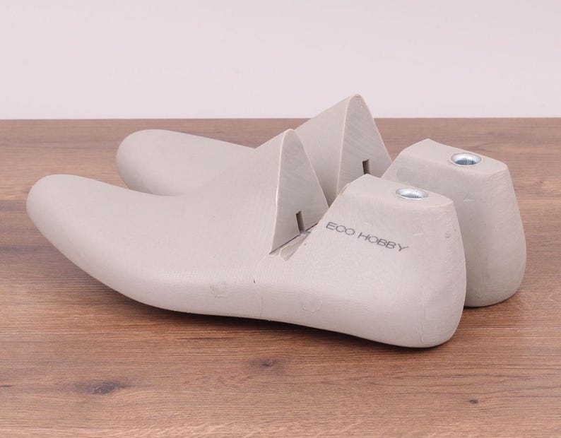 May include: Two light gray shoe lasts, one with the text "ECO HOBBY" printed on the side, are displayed on a wooden surface. The lasts are designed to maintain the shape of shoes during storage or repair.