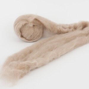 May include: A close-up shot of a light brown, fluffy fiber material, likely wool or a similar textile. The material is rolled into a compact ball at one end, with the rest of the fiber extending outwards. The texture appears soft and delicate.