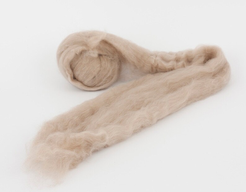 May include: A close-up shot of a light brown, fluffy fiber material, likely wool or a similar textile. The material is rolled into a compact ball at one end, with the rest of the fiber extending outwards. The texture appears soft and delicate.