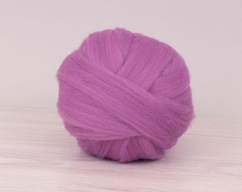 Puede incluir: Una bola de vellón de lana morada vibrante, bien enrollada y texturizada. La lana es de color liso, con un aspecto suave y esponjoso. El fondo es una superficie clara con vetas de madera.