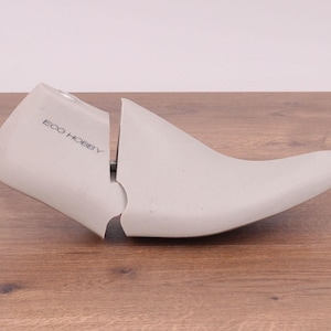 May include: A light gray shoe stretcher with the text "ECO HOBBY" printed on the side. The shoe form is made of a solid material and is split into two pieces. The shoe stretcher is resting on a wooden surface.