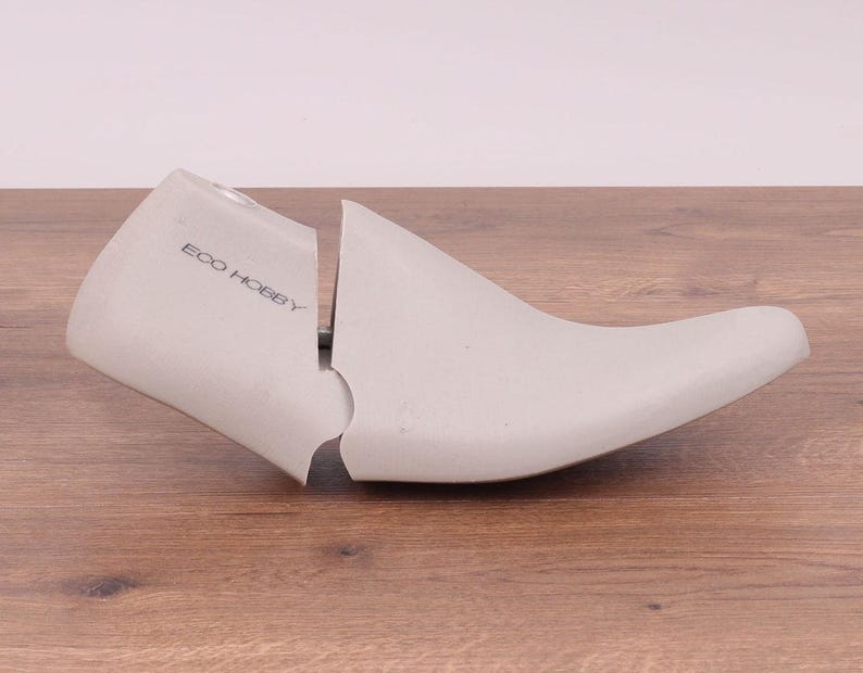 May include: A light gray shoe stretcher with the text "ECO HOBBY" printed on the side. The shoe form is made of a solid material and is split into two pieces. The shoe stretcher is resting on a wooden surface.