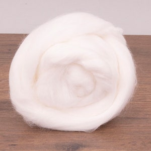 May include: A large, fluffy ball of white wool roving, ideal for fiber art projects. The wool is tightly packed and appears soft and cloud-like. The background is a light wood surface.