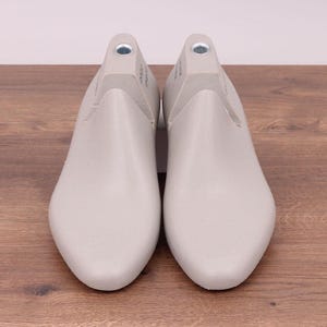 May include: A pair of light gray shoe lasts, designed for shaping footwear. The lasts are made of a smooth material and are positioned on a wooden surface. The shoe forms have a rounded toe and a heel area with a metal insert.