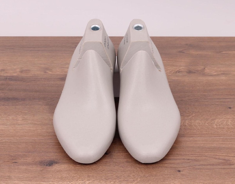May include: A pair of light gray shoe lasts, designed for shaping footwear. The lasts are made of a smooth material and are positioned on a wooden surface. The shoe forms have a rounded toe and a heel area with a metal insert.