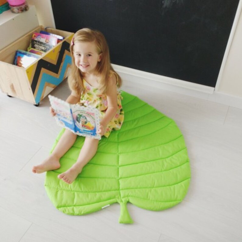 May include: A young girl sits on a large green leaf-shaped play mat reading a book. The mat has a quilted texture and is shaped like a large leaf.