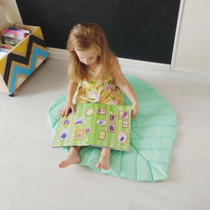 May include: A young girl sits on a light green, quilted, leaf-shaped play mat and reads a book with colorful illustrations.