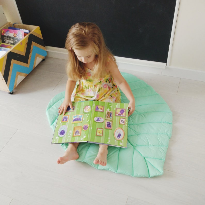 May include: A young girl sits on a light green, quilted, leaf-shaped play mat and reads a book with colorful illustrations.