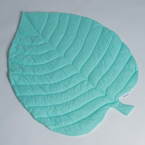 May include: A light green quilted leaf-shaped play mat. The mat is made of soft fabric and has a quilted design that resembles the veins of a leaf.