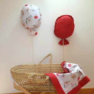 May include: A white wicker bassinet with a red and white floral patterned blanket and two fabric balloons hanging on the wall behind it. One balloon is red and the other is white with red floral accents.