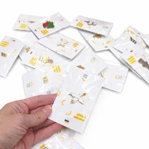 Könnte beinhalten: Eine Sammlung von verpackten Anstecknadeln. Eine Nadel zeigt eine goldfarbene Taube mit einem Olivenzweig. Die Verpackung trägt den Text "elvi me" und eine ukrainische Flagge. Weitere Anstecknadeln haben verschiedene Designs.