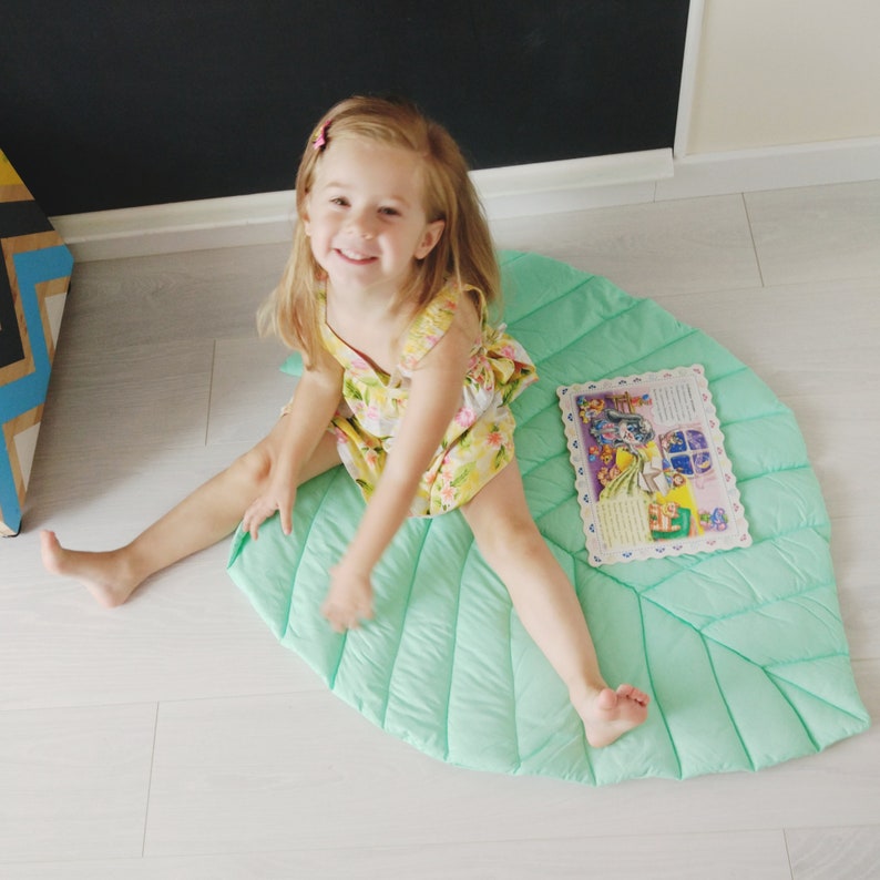 May include: A young girl sits on a large, green, leaf-shaped play mat. The mat has a quilted texture and is surrounded by a white wooden floor. The girl is wearing a floral dress and is smiling at the camera. A book with colorful illustrations is lying on the mat.