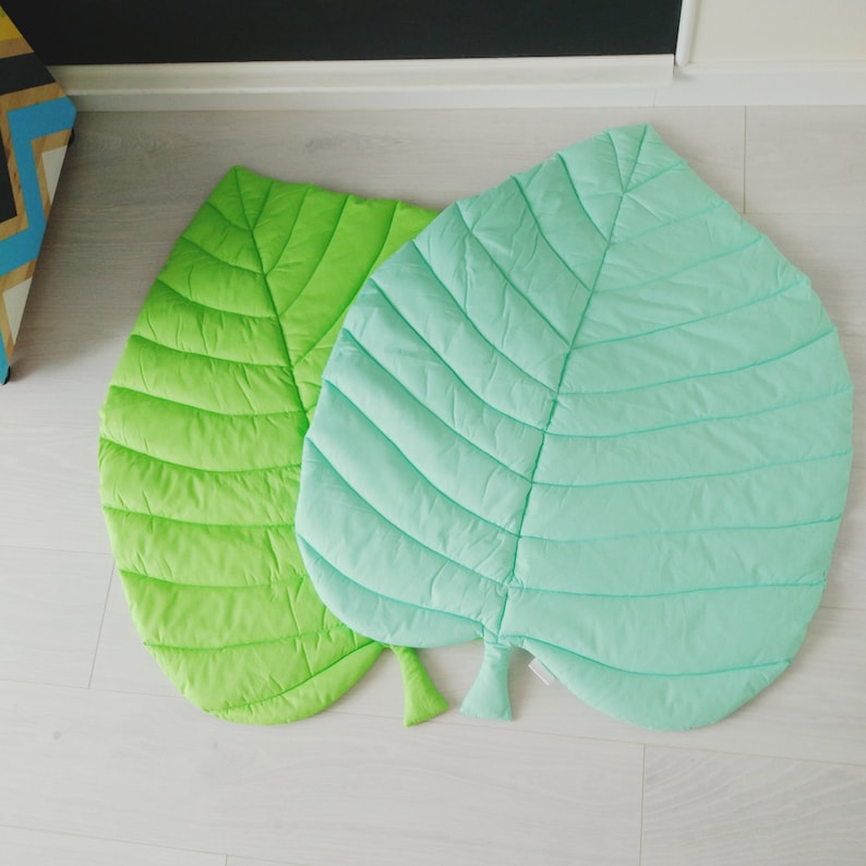 May include: Two quilted leaf-shaped play mats, one green and one light blue. The mats are made of soft fabric and have a quilted design.