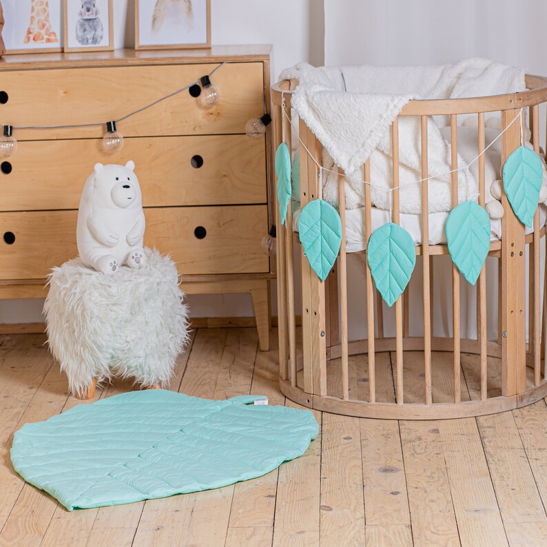 May include: A light green leaf-shaped play mat for babies and toddlers. The mat is on a wooden floor next to a crib with a white blanket and a garland of light green leaves.