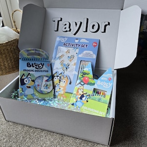 Pode incluir: Uma caixa de presente branca cheia de itens de atividades com o tema Bluey. Inclui um bloco de aquarela mágica, um conjunto de atividades e um conjunto de colorir. A caixa é personalizada com o nome "Taylor".