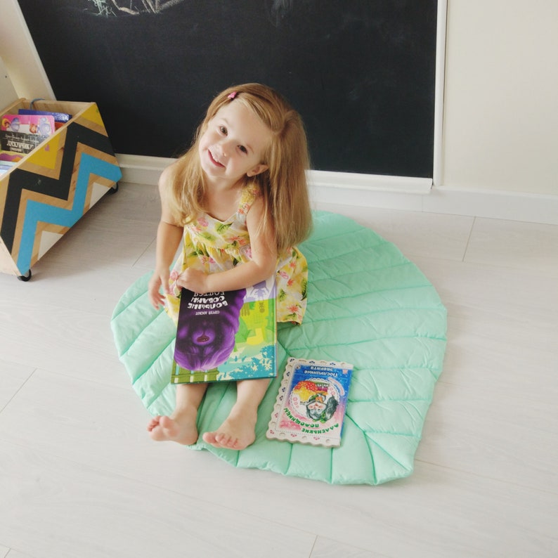 May include: A young girl sits on a light green, quilted play mat with a book titled "The Very Busy Spider" in her hands. She is wearing a yellow floral dress and has long blonde hair.