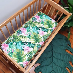 May include: A wooden crib with a white fitted sheet featuring a tropical print of pink flamingos and green palm leaves. The crib is sitting on a green leaf-shaped rug.